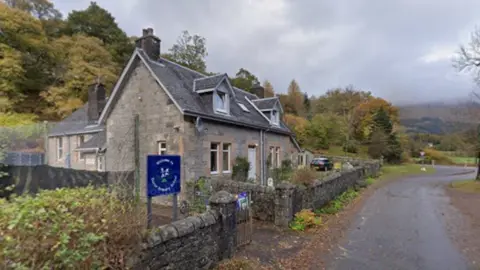 The school is a Victorian era-style building. There is a blue sign for the school and trees behind it.