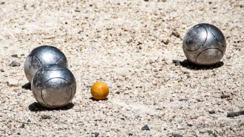 Three metal Pétanque boules sit apart from eachother on a gravel floor, all three are shining in the sunlight.