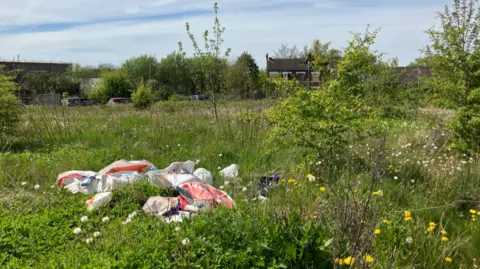 Bags of waste lie on overgrown grass and weeds, with derelict buildings in the distance