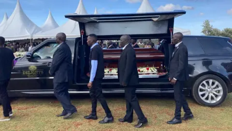 Anne Soy/BBC Men walking by a hearse