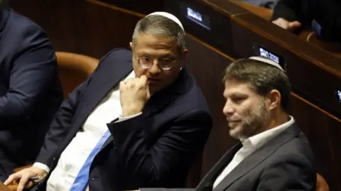 Itamar Ben-Gvir, left, a man with grey hair wearing glasses and a white cap, sits beside Bezalel Smotrich, a man with dark hair and greyish beard, wearing a white cap.