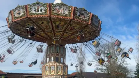 BBC/ Emily Johnson A colourful swing ride spinning with riders lifted outward on chains, set against leafless trees, red‑roofed buildings and a bright blue sky with scattered clouds.