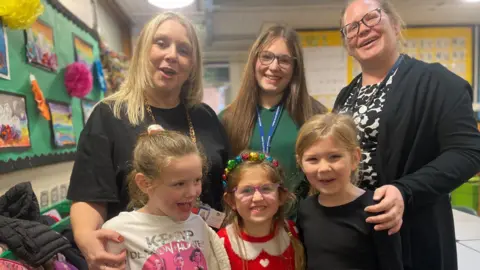 Simon Thake/BBC Three small children in party clothes smile at the camera. Behind them an older girl in a green sweatshirt is flanked either side by two adults who put their hands of the children's shoulders.