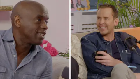Trevor Nelson wearing a light grey shirt and Scott Mills sat on an sofa wearing a denim jacket.