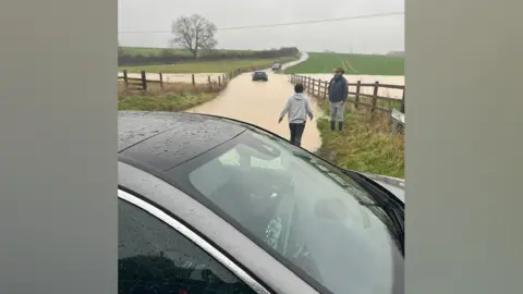 Helen Louise Brown Cars in floodwater on a rural road. Some people on foot are in attendance.