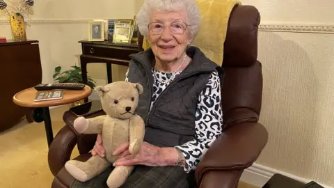 BBC Gabriele Keenaghan sitting in a chair with a teddy bear on her lap. It was the bear she brought with her from Vienna on the Kindertransport