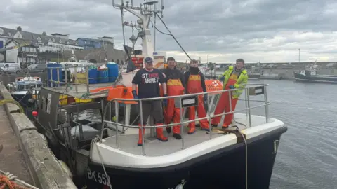Four men are standing on a boat with Seahouses harbour behind them. They are all wearing waterproofs 