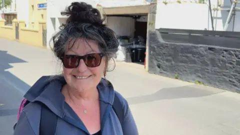 Mandy is smiling at the camera as she stands on a pavement on a lane. She is wearing sunglasses and a small rucksack. Her dark and grey hair is pinned into a top knot. 