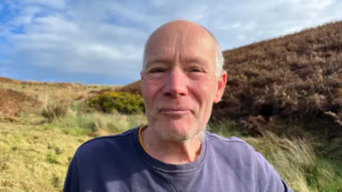 Rob is standing on a hill, with green and brown grass and marsh in the background. He is wearing a navy sweater. He has very short white hair and is smiling at the camera