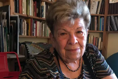 A woman looking at the camera with grey hair and a bookcase behind her