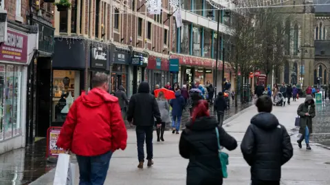 Jacob King/PA Wire Shoppers with shopping bags, walking down a busy high street. Some seasonal festive lights can also be seen. 