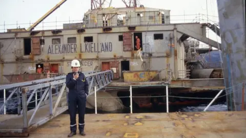 Sven Tønnessen/Norwegian Petroleum Museum Old picture of a man in overalls and a hard hat standing in front of the wreck of the Kielland