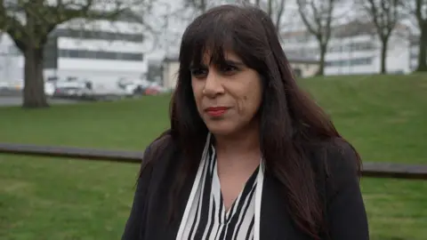 Woman with dark hair and a fringe, wearing a striped blouse and a black blouse, stood in front of a field with white buildings in the distance 
