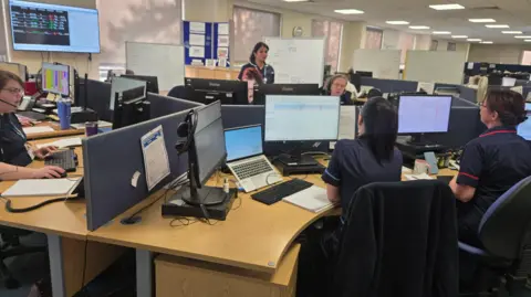 A group of women in nurses' uniforms sit at computers in an office. A number of whiteboards are in the background.