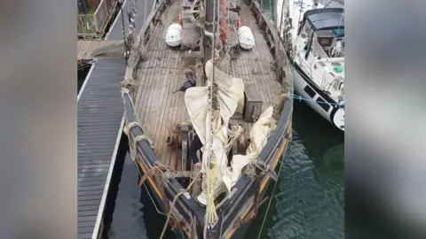 Jamie Niblock/BBC An aerial of the front of the Nordlys ship. The outside of the ship is painted black and the top deck is made from dark wood. The boat is moored on water and is sitting next to a white boat.
