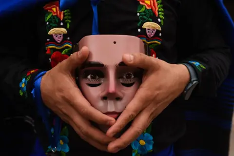 Hector Quintanar/Getty Images A close-up view  of a persons hands holding the traditional mask of Parachico - a man's face with black eye make-up.