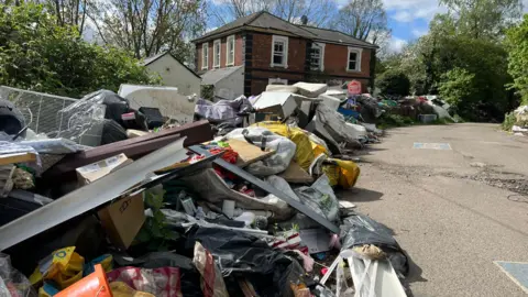 The old station, now with broken and boarded up windows. All along the road in front of it, and around the building, is flytipping - mattresses, white goods, blankets, and all kinds of rubbish. There is a red For Sale sign in front.