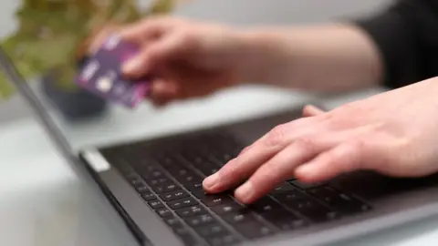 PA Media A woman using a laptop as she holds a bank card