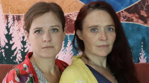 Donald Sage Mackay Penelope, with light brown hair, and Ginny Skinner, with deep red hair, both facing towards the camera