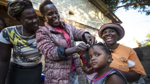 Getty Images Woman plaits a little girl's hair whilst two other women look on