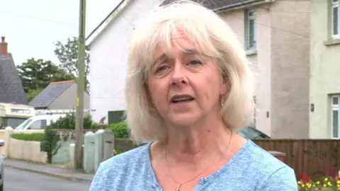 Ruth Jones standing on a street with houses and a pavement in the background. She has white shoulder length hair and is wearing a blue shirt and a necklace with a small cross 