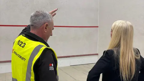 South Ribble Borough Council handout A builder (left) with grey hair and wearing a hi-vis vest over a black long-sleeved top discussing the building work with councillor Clare Hunter who has long blond hair and wear a black blazer jacket. The image is taken from behind. They are standing in an old squash court.