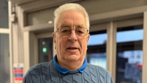 Robert Constantin/ BBC A man with white hair and glasses in a blue polo and a grey cable knit jumper stands central to the camera. 