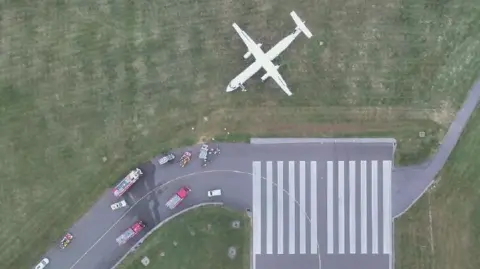An aerial view of the white aircraft on the grass area of the runway it is facing horizontally compared to the direction of the runway itself. Multiple vehicles are seen parked on the runway.