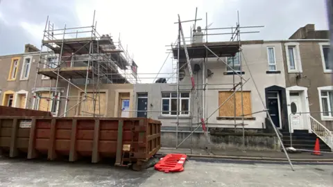 A row of two-storey terraced houses has a large upper floor section missing. Scaffolding surrounds the structure.