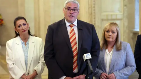 PA Media A formal indoor setting with two women and a man standing close together behind a microphone. The central person, Gavin Robinson, wears a dark suit with a white shirt and a diagonally striped orange and red tie and appears to be speaking. On either side of the central speaker stands Emma Little Pengelly and Michelle McIlveen in light-colored jackets, facing forward. A microphone with a small logo panel is positioned in front of the speaker. The background shows an ornate interior at Stormont with marble walls.