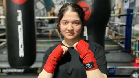 Shariqua Ahmed/BBC Nazia wearing a black T-shirt and red bandages around her wrists. She is holding her fists up to her chin posing for the camera. A boxing ring is behind her.