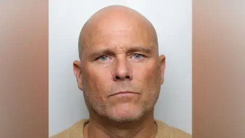 Mugshot of Richard Whitaker, who has a bald head, stubble and is wearing a yellow jumper.