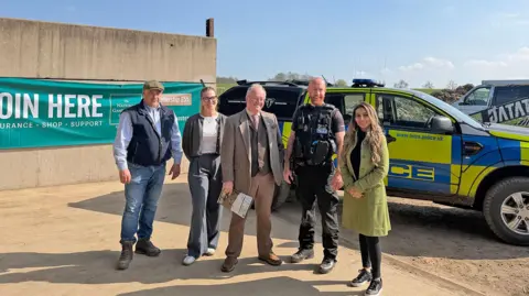 Office for the Leicestershire and Rutland police and crime commissioner Five people stand in front of a police car smiling. It is a sunny day. 