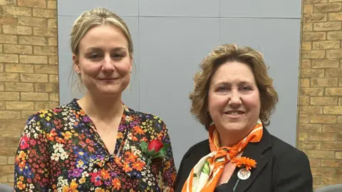 Two women, on the left Deputy Leader Lauren Townsend, and on the right Leader Jane Carr, stand side by side indoors in a formal setting, with a UK flag displayed behind them against a brick wall.