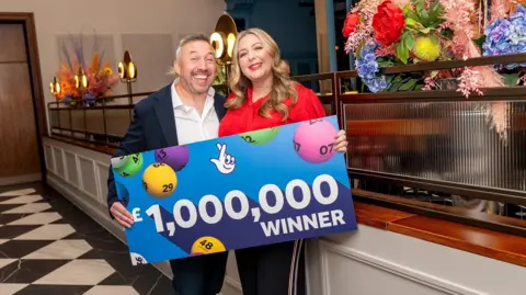 National Lottery A man and a woman hold up a large blue card with the words '£1,000,000 winner' written on it.