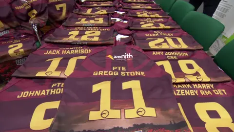 A table full of maroon rugby shirts bearing the names of armed forces veterans above large numbers.