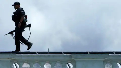 Armed Secret Service agent walks right to left along the top of the North Portico of the White House