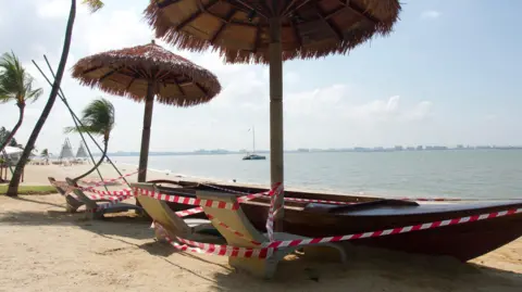 BBC/ Gavin Butler A wooden boat styled into a bench on a beach, under palm-leaf umbrellas and alongside stone banana chairs, wrapped in red-and-white "caution" tape. Ocean in the background, and on the distant horizon is a faint skyline of buildings.
