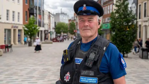 Allan stands on a pedestrianised street with shops either side, facing the camera. Blurred in the background are a few shoppers milling about but the street is quite empty. He wears a blue t-shirt under a back police vest, which carries two walkie-talkies and has the Essex Police emblem on. A blue label on his vest says Police Community Support Officer. He wears a black police hat with royal blue riband. He is clean shaven and has an earpiece on his left ear.