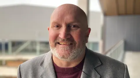 BBC A man smiles toward the camera, he is wearing a burgundy jumper and grey suit jacket. He has a short grey beard, and bald head.