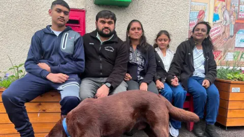 The Patel family are pictured sitting outside a post office with their dog. A letter box can be seen in the wall behind them and there is also a picture on the wall. Umang is second on the left. He is sitting beside a teenage boy who is wearing a dark blue tracksuit. On the other side of Umang is a woman and she is sitting next to a young girl and another woman. The dog is brown.