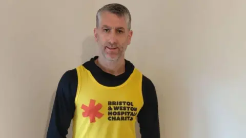 A man in his 40s with grey hair stands against a white wall wearing a black hoodie with a yellow Bristol and Weston Hospitals Charity vest top on.