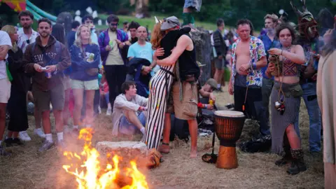 Ben Birchall/PA Media Two people hugging next to a fire at Glastonbury, surrounded by other people.