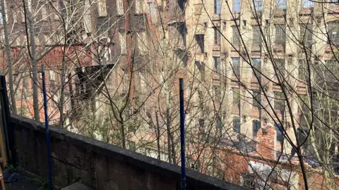 A six-storey mill building seen from a nearby rooftop, behind tree branches.