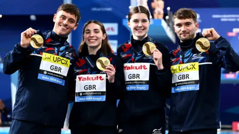 Getty Images Gold Medalists, Daniel Goodfellow, Thomas Daley, Scarlett Mew Jensen and Andrea Spendolini Sirieix of Team Great Britain pose with their medals during the Medal Ceremony after the Mixed Team Event Final on day one of the Doha 2024 World Aquatics Championships. The team all wear navy blue British Swimming tracksuits and smile at the camera.