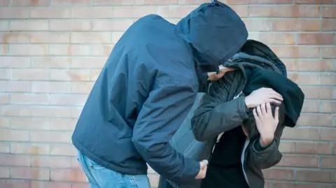 This is a stock photograph depicting two young men wearing dark, hooded clothing. One is holding the shoulder of the other in an aggressive stance, with the other fist clenched. The other is cowering and has his hands protecting his head.