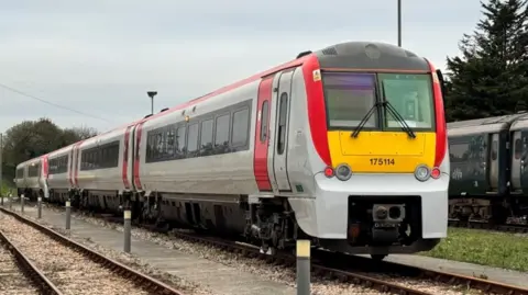 GWR A 4-carriage long grey train with red doors in a siding next to a dark green train and surrounded by trees.