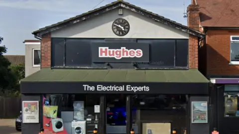 Exterior of a Hughes shopfront. Washing machines and other white appliances can be seen in the shop window. Written on a green awning is the phrase, the electrical experts.