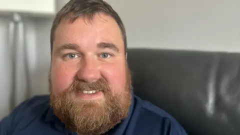 Kevin Clemitson is sitting on a black leather couch. He has mid brown hair and a lustrous beard and moustache and is fair skinned with freckles. He is smiling and wearing a navy blue top. 