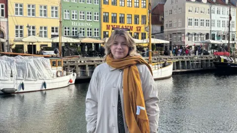 Annabelle is stood in front of coloured houses by a canal. Boats a visible. She is wearing an orange scarf and a white coat. 